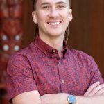 man with patterned shirt and dreads with māori carving in background